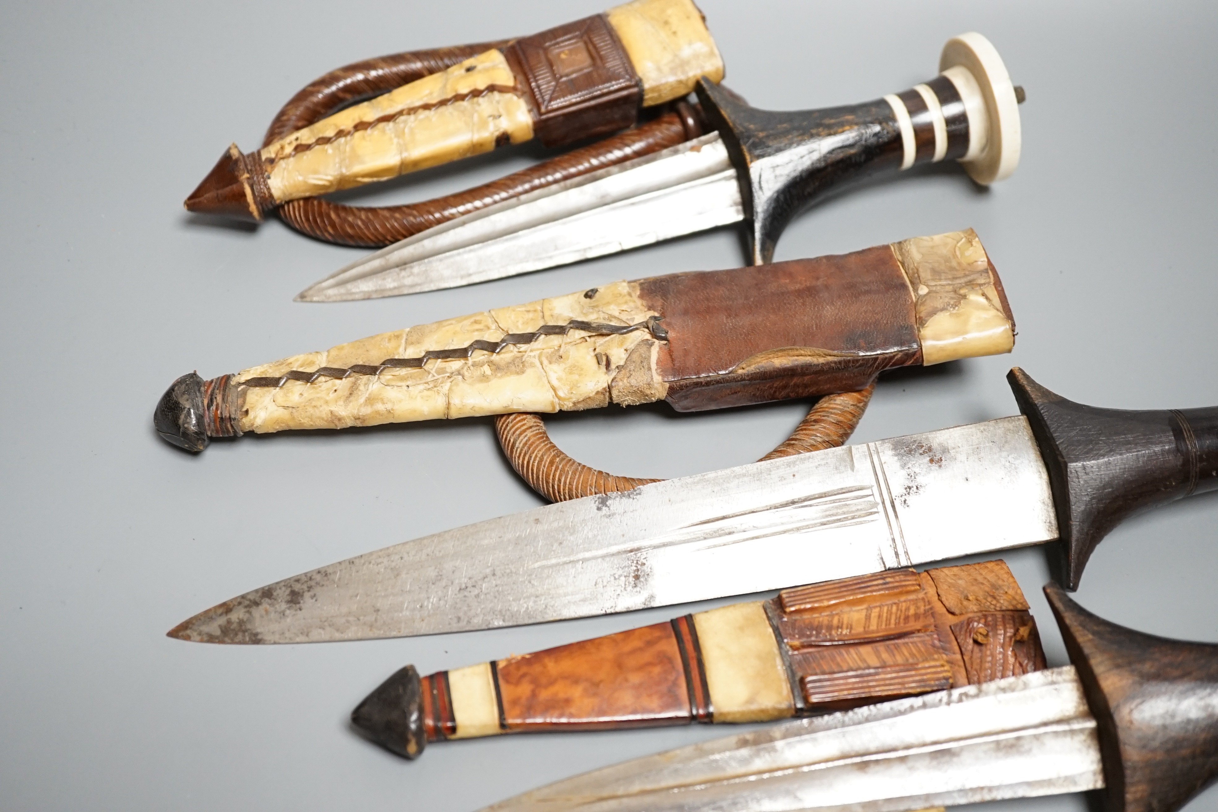 Five assorted African Nilotic tribal daggers c.1900, comprising three with turned ivory mounted ebony handles (two with silver pommels); and two further with cylindrical ivory handles, all in their leather sheaths, maxim